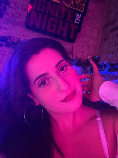 A person with dark hair is lying down, their face illuminated with pink lighting. They have fair skin, dark eyes, and are wearing hoop earrings. Their hand is near some foliage lit with blue light. A sign that reads "Ignite the Night" is in the background, partially obscured. The location is unknown.