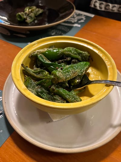 A yellow bowl filled with sautéed green peppers sits on a gray plate. A silver spoon is buried within the peppers. A smaller, dark bowl also contains peppers. A wooden table serves as the backdrop, partially covered by coasters or menus with green, white, and black designs.