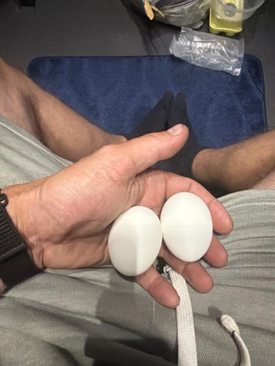 The image shows a person holding two white, oval objects in their right hand. The objects appear to be eggs. The person is wearing grey sweatpants and a black watch on their left wrist. In the background, a blue mat and a person's feet wearing black socks are visible. There are also some plastic bags and a yellow bottle in the background. The scene appears to be indoors, possibly in a living room or bedroom. The location in the city of New Britain, United States of America is not specified.