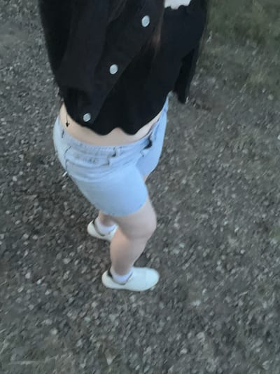 A low-angle, high-contrast, vertical shot shows a person's legs and torso from the waist down, walking on a coarse, grey, asphalt surface. The person is wearing light blue denim shorts, a black jacket with silver buttons, and white sneakers with white socks. The scene appears to be outdoors, possibly during the daytime due to the lighting. The setting seems to be a road or pathway. There are no visible text elements in the image.