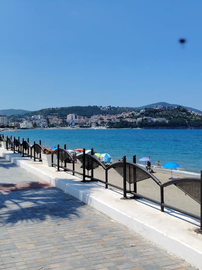 The image shows a view of the coastline in Agropoli, Italy, on a clear, sunny day. In the foreground, a paved walkway runs along a white-painted concrete barrier topped with a black metal fence. Beyond the fence is a sandy beach with several beach umbrellas and a few people visible. The sea is a vibrant blue, and in the background, the city of Agropoli stretches along a hilly coastline, with buildings and vegetation covering the slopes. The sky is a clear, bright blue, with a small, blurry dark spot near the top center of the image, possibly an artifact or insect. The overall scene depicts a typical summer day at a coastal resort town.
