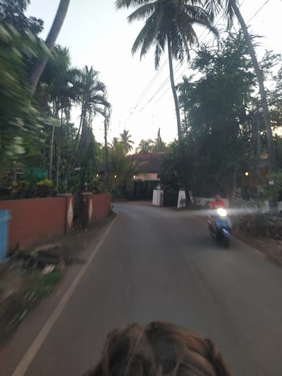The image depicts a road in what appears to be a tropical area, likely near Marna, India, given the presence of coconut trees. A scooter with its headlight on is moving along the road. The scene appears to be taken from a moving vehicle, judging by the motion blur. A section of a brick wall runs along the edge of the road.