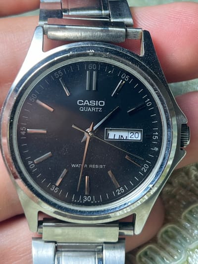 A close-up shot shows a black analog-digital Casio watch with a metal band. The watch face features hour markers and numbers along the bezel, with the brand name "CASIO QUARTZ" displayed at the top of the face. A digital window at the 5 o'clock position shows "LUN 20", indicating Monday the 20th. The watch is held by a hand, partially obscuring the upper left and right corners of the frame. The background is out of focus and appears to be a textured green surface, possibly fabric or a plant. The scene does not depict any identifiable urban or natural landmarks in Romitan, Uzbekistan.