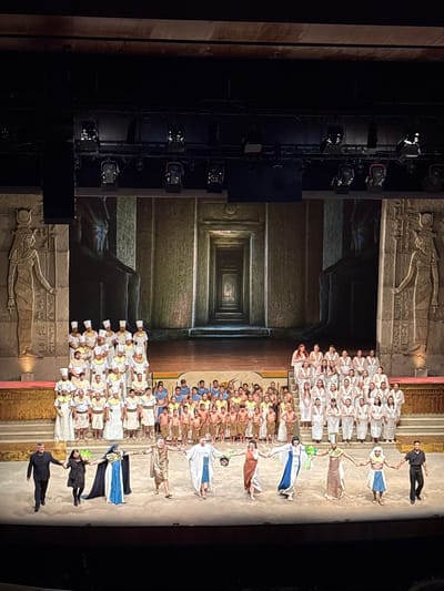 A stage performance is taking place in San Borja, Peru. The backdrop is an image of an Egyptian tomb. The performers include several men and women wearing Ancient Egyptian-style robes. Some are dressed in white robes, while others are in brown and blue. The foreground shows some black equipment and spotlights. Two men dressed in black appear to be managing the performance.