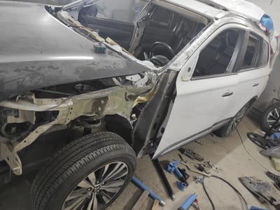 The image shows a white SUV, likely undergoing repairs in a workshop. The front fender and hood are open, revealing internal mechanics and damaged bodywork. Various tools and parts are scattered on the concrete floor around the vehicle, including blue pneumatic tools, pliers, and metal plates. The interior of the car is visible, with black seats and a steering wheel. The overall environment suggests a car repair or body shop. The city of Qairat, Kazakhstan is mentioned as the location, but there are no visual cues in the image to confirm this specific location.
