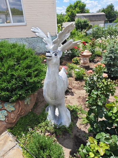 A concrete statue of a deer stands in a garden in Krutinskiy, Russia. The deer statue is painted silver and has antlers, a long neck, and large eyes. It is positioned in the center of the frame, with a lush green bush to its left and a rose bush to its right. In the background, there is a garden with various flowers, including roses, and two small buildings. The scene appears to be set on a sunny day, with some clouds in the sky. The overall impression is one of a peaceful, well-maintained garden with decorative elements. There are no people visible in the image, and no text is present.