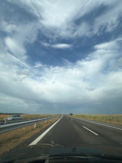 The image shows a highway scene in Zheleznik, Bulgaria, taken from inside a vehicle. The perspective is from the driver's side, looking forward and slightly to the left. A highway stretches out into the distance, with clear white lane markings on the dark asphalt. A metal guardrail runs along the right side of the road, separating it from a dry, grassy field. The field is dotted with small white flowers, suggesting a rural or steppe-like environment.

The sky above is a dramatic mix of blue and white clouds. Wispy, feathery clouds are interspersed with larger, more solid formations, indicating a dynamic weather pattern. The lighting suggests it is daytime, likely late morning or afternoon, with the sun partially obscured by clouds.

A silver car is visible parked on the shoulder of the road, behind the guardrail. The overall mood of the image is one of travel and open road. There are no people clearly visible. The scene is tranquil and expansive, capturing a moment of transit through the Bulgarian countryside.