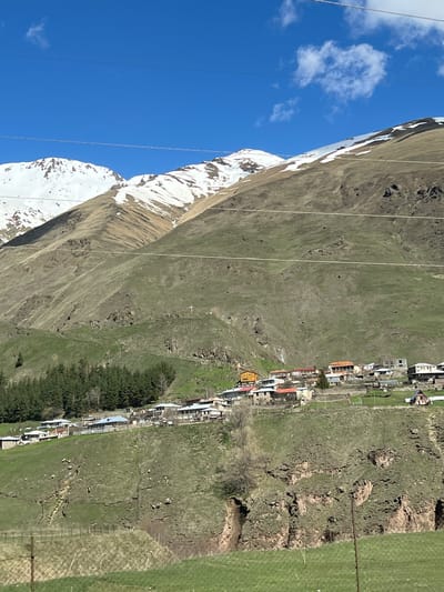 The image shows a landscape in Sioni, Georgia. A snow-capped mountain occupies the left side of the frame, contrasted against a clear blue sky with scattered white clouds. Power lines are visible in the sky. A village is situated at the base of the mountain, interspersed with green fields and patches of forest. The landscape appears rugged and mountainous.
