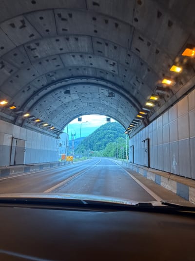 The image shows a perspective from inside a vehicle exiting a tunnel. The tunnel ceiling is constructed from concrete segments, with regularly spaced lights illuminating the interior. The walls of the tunnel are paneled and appear to be made of metal or a composite material. Orange lights are visible on the right side of the tunnel, suggesting illumination. The road surface is wet, indicating recent rain or a damp environment.
As the vehicle approaches the tunnel exit, the scene transitions to an exterior view. Lush green mountains covered in trees dominate the background, with a bright, cloudy sky overhead. A paved road curves to the right, with a white line marking the lane division. Various structures are visible near the tunnel exit, including what appears to be a traffic signal pole with red and white indicators, a yellow barrier, and some industrial-looking buildings or access points along the tunnel wall. A sign with a red circle and a diagonal line, likely indicating a prohibition, is visible on the left side of the road.
The image does not contain any people or explicit actions. The focus is on the infrastructure of the tunnel and the natural landscape at its exit. The location identified is the Kepsha Tunnel in Kepsha, Russia.