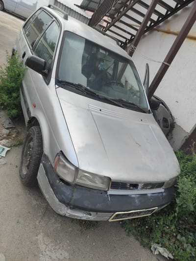 A silver station wagon is parked on a dirt and gravel lot next to a building with a metal awning. The car is in disrepair, with a dented front bumper, cracked paint on the hood, and overgrown vegetation encroaching on its sides. The driver's side door is ajar. The scene appears to be in an industrial or junkyard area in Qairat, Kazakhstan.