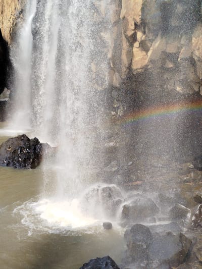 The image depicts a waterfall. The water cascades down rocks, creating a fine spray in the air. A rainbow is visible in the mist near the right side of the image. The waterfall empties into a pool of water surrounded by rocks.