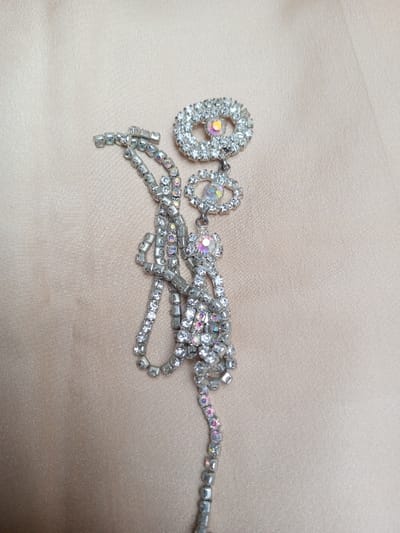 The image displays a single piece of costume jewelry, likely a brooch or decorative element, positioned on a smooth, light beige or champagne-colored fabric surface. The jewelry item is composed of silver-toned metal set with numerous clear and iridescent rhinestones.
The upper portion features two interconnected, rhinestone-encrusted oval elements. The top-most oval is larger, oriented horizontally, and contains a central, round iridescent rhinestone. Directly below it, a smaller, horizontally-oriented oval is suspended, also containing a central, round iridescent rhinestone.
Attached to the bottom of the smaller oval is a multi-strand tassel-like dangle. This dangle originates from a central, round iridescent rhinestone encased in a smaller rhinestone-studded setting, from which multiple chains of clear rhinestones cascade downwards. These chains vary in length and arrangement, creating a clustered effect. The entire piece is seen from a slightly elevated, oblique angle, with the jewelry predominantly positioned in the upper right and central-right portions of the frame, extending downwards. No people, actions, or interactions are present in the scene. The implied location is Qarabucaq qəsəbə, Azerbaijan.