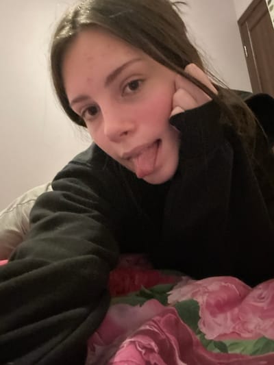 A young woman in a black hoodie playfully sticks out her tongue. Her dark hair frames her face, and her left hand is gently touching her cheek. The background is a mix of soft textures, including what appears to be a floral-patterned blanket or bedding in pink and green hues. A wooden cabinet is visible in the top left corner of the image. The photo is taken from a slightly angled, close-up perspective.