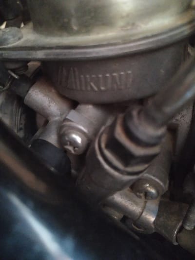 The image is a close-up, slightly blurred photograph of a carburetor from a motorcycle or similar engine, likely located in Gampongbaro, Indonesia. The carburetor is made of metal and has various components visible, including screws, bolts, and hoses. There is a raised logo or text on the carburetor that appears to read "MIKUNI." The surrounding environment is dark and appears to be the engine bay of a vehicle. No people or other living subjects are visible.
