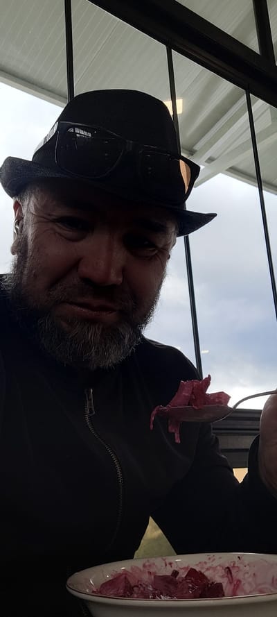 A medium shot captures a man wearing a black fedora and sunglasses, with a salt-and-pepper beard, seated indoors and looking towards the camera. He is holding a spoon filled with what appears to be chopped beets, poised to eat from a bowl of similar contents. The man is dressed in a dark jacket with a visible zipper. The background features a modern interior with white slatted ceilings and large windows revealing an overcast sky. The scene is set in a restaurant in Milisi, Georgia.