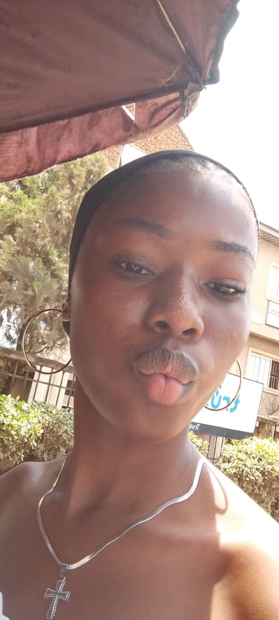 A medium-shot portrait captures a dark-skinned individual, appearing female and young, positioned outdoors in Ohokobe Ndume, Nigeria. The subject's head is slightly tilted, with eyes partially closed and lips pursed into a "kissing" expression. She wears a dark, possibly black, headband and large, circular, metallic hoop earrings. A silver-colored, flat link chain necklace with a prominent cross pendant rests on her exposed upper chest and collarbones. Bright sunlight illuminates the scene from above, casting some definition across the subject's features. Above the subject's head, a section of a reddish-brown, faded fabric canopy or umbrella is visible, showing signs of wear. The background includes green foliage, suggesting bushes or trees, and to the right, a light-colored building with dark window frames and white security grilles. A partially visible white vertical sign with blue or black lettering, reading 'TRUST' at the bottom and part of 'HELPERS' at the top, stands in the mid-ground.