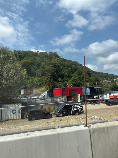 The image depicts an outdoor scene in Vysokoye, Russia, characterized by a collection of shipping containers repurposed into structures. In the foreground, a concrete barrier partially obscures the view. Behind it, a gravel-covered area is visible with several parked vehicles, including a black van with "ENDURO 23" branding and a black sedan. Various shipping containers are stacked and arranged to form buildings, some painted red and blue. One container has "ENDURO 23" branding, and a yellow ATV is parked on top of a red container. A densely wooded hill forms the backdrop. The sky is partly cloudy.