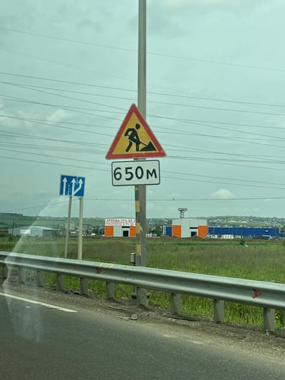 The image shows a roadside scene with several traffic signs in Solontsy, Russia. In the foreground, a road with a metal guardrail runs along the right side. Beyond the guardrail is a grassy field. In the background, there are buildings, including some under construction with orange and white panels, and a city skyline. The sky is overcast.
The traffic signs are mounted on poles. The topmost sign is a yellow triangle with a red border, depicting a person with a shovel, indicating road work ahead. Next to it is a rectangular sign with the number "650" in black on a white background, likely indicating the distance to the road work. Below these, a blue rectangular sign shows two lanes merging into one, with arrows indicating traffic should move to the right.
A smaller sign with text is visible on a pole in the background, appearing to be an advertisement. It reads "АРЕНДА 214-44-44" and "И СТРОИТЕЛЬСТВО 271-22-1". This suggests the rental and construction business.
The scene appears to be captured during the daytime, with the cloudy weather creating a diffused light. The overall impression is a functional roadside environment with ongoing construction or maintenance work.