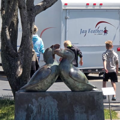 A bronze sculpture of two puffins, with their heads facing each other, is located on a stone pedestal in front of a white trailer with the text "Jay Feather" and a cartoon bird logo. In the background, a tree trunk is visible on the left. Three people are present: a woman wearing a light blue hooded sweatshirt, a boy in a black and green hooded sweatshirt, and another boy in a camouflage hooded sweatshirt and black shorts. The scene is outdoors on a sunny day, with grass and asphalt visible in the foreground and background, respectively. The image appears to have been taken in Cannon Beach, Oregon, United States of America, known for its distinctive rock formations and puffin populations.