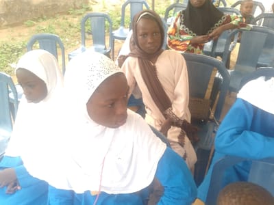 This image depicts a daytime gathering in Ijoko, Nigeria. Multiple individuals, primarily children and young women, are seated on blue plastic chairs in what appears to be an outdoor or semi-sheltered area with an earthen ground.
In the foreground on the left, two young girls are seated. The leftmost girl is partially obscured, wearing a white hijab and a blue garment, looking downwards. To her right, a second girl, also in a white hijab (adorned with small dark embellishments) and a blue top, looks towards the right. On the right side of the foreground, the upper body of another individual in a white hijab and blue garment is partially visible.
In the mid-ground, slightly to the left of center, a young girl with a light brown hijab and a long-sleeved, light-colored dress is seated, looking directly at the viewer with a neutral expression. Her arms are crossed in her lap, and her sleeves feature patterned cuffs. Behind her, on a blue plastic chair, a dark brown, patterned handbag is visible. Further back and to the right, an adult woman in a black hijab and a colorful, long-sleeved patterned dress is partially visible, leaning forward and to the right, with her hands clasped. A small child's head is partially visible in the far background on the right.
Numerous empty blue plastic chairs are scattered in the background, suggesting a larger seating arrangement. A light-colored wall or structure is visible in the upper left background.