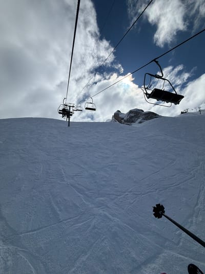 The image shows a ski slope in La Giettaz, France. A ski lift runs along the left side of the frame, with several empty chairs visible against a cloudy sky. The slope itself is covered in snow, with visible tracks indicating skier activity. A ski pole is partially visible on the right side of the image. The general environment suggests a winter sports setting in a mountainous region.