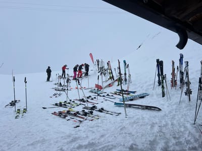 The image depicts a snow-covered mountain slope at a ski resort in Valtournenche, Italy, under an overcast sky with visible precipitation. Six individuals in full ski attire are present in the mid-ground. To the far left, one person in a black jacket and dark pants stands, facing right, holding ski poles. To their right, five additional individuals are grouped, dressed in colored ski jackets and pants (red, pink, dark grey), primarily bent over, appearing to adjust ski equipment. The foreground and mid-ground are densely populated with multiple sets of skis and ski poles, both lying flat on the snow and standing upright, stuck into the snow. At least two snowboards are visible on the snow, one clearly marked "WHATEVER." A red triangular banner stands upright on the far right. An architectural overhang, likely part of a building structure with a dark gutter, is visible in the upper right quadrant of the frame.