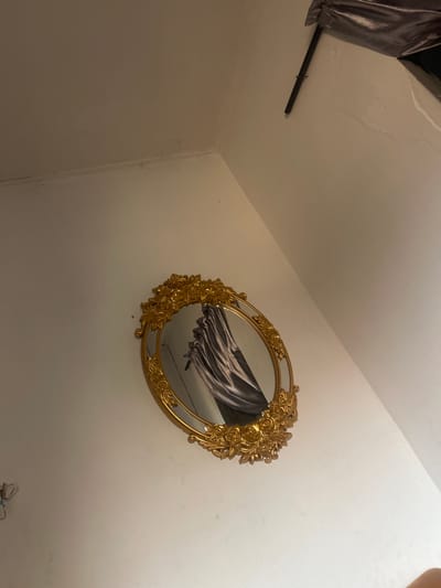 The image depicts an indoor scene in Ilishan, Nigeria, focusing on an oval-shaped mirror with a prominent gold, ornate frame featuring floral motifs, mounted on a plain white wall. The mirror's reflection shows a textured, dark grey or silver fabric, consistent with a drape or curtain, partially obscuring a dark background. The wall surface appears smooth, with a subtle seam or corner visible in the upper left. In the upper right corner of the overall scene, a dark, thin rod is horizontally mounted, partially obscured by a section of dark, metallic-sheen fabric, possibly part of an awning or another decorative element, that is slightly angled downwards from the ceiling. No people are visible. The environment suggests an interior space with minimal furnishings beyond the mirror and the reflected elements.