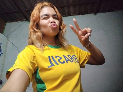 A young woman with auburn hair poses for a selfie. She is wearing a yellow t-shirt with green accents, and the word "BRASIL" is printed in blue across the chest. Her hair is styled in loose waves, and she has a playful expression, puckering her lips and winking at the camera. Her right hand is raised, forming a peace sign. The background appears to be an indoor setting, possibly a room with a textured wall and a wooden ceiling. The lighting is dim, suggesting it could be evening or nighttime. There are some wires visible on the wall. The woman is wearing a bracelet on her right wrist and has a tattoo on her inner forearm. The overall mood is casual and cheerful.
