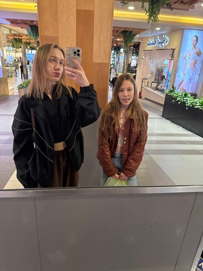 A selfie taken in a mirror shows two women. The woman holding the phone has blonde hair and is wearing a black jacket and brown top. The other woman, reflected in the mirror, has long brown hair and is wearing a brown jacket and jeans. They are standing in a shopping mall, as evidenced by the stores and decorations visible in the background. The exact location in Ol'ginskaya, Russia is not discernible from the image alone.