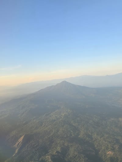 An aerial view captures a majestic, cone-shaped mountain peak rising from a vast, verdant landscape. The mountain is covered in dense green foliage, with a prominent ridge extending from its base. The surrounding terrain is also densely forested, with rolling hills and valleys stretching towards the horizon. In the distance, a hazy layer of mist softens the outline of more distant mountain ranges. The sky above is a clear, pale blue, with a faint band of warmer, lighter hues near the horizon, suggesting either sunrise or sunset. The overall scene is one of natural beauty and vastness. The location is Ujongkeupula, Indonesia.