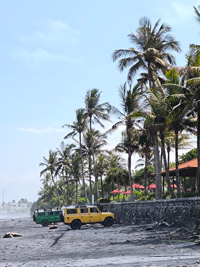 The image shows a beach scene in Selemadeg Timur, Indonesia. Two vintage Land Rover vehicles, one green and one yellow, are parked on a black sand beach. A row of palm trees lines the back of the beach, along with a low stone wall and a building with a red tile roof. In the background, there is a structure resembling a tower and several buildings. The sky is a pale blue with some clouds. Debris is scattered on the beach, particularly near the stone wall.