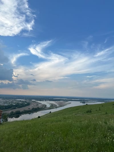 The image shows a scenic view of a wide river valley from a grassy hilltop. The sky is a vibrant blue with streaks of wispy white clouds. The foreground is dominated by a lush green meadow dotted with small white flowers. A portion of a reddish-brown brick structure is visible on the far left of the foreground. The mid-ground features the winding river, flanked by green trees and patches of land. In the distance, a flat landscape stretches out, with hints of human settlement and a bridge crossing the river. The overall scene evokes a sense of natural beauty and tranquility. This view is characteristic of the landscape in Britvino, Russia, known for its rolling hills and riverine scenery.