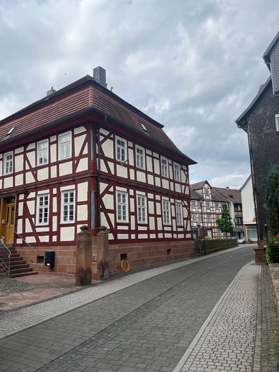 The image depicts a street scene in Neustadt, Germany. The foreground features the side of a traditional half-timbered building with dark red timber framing and white infill. Numerous white-framed windows are visible on this building. A paved sidewalk runs alongside the building, composed of grey and lighter grey cobblestones. Further down the street, another half-timbered building, with a similar architectural style, is visible, along with a tree. The overall setting appears to be a town or village.

The sky is overcast with heavy clouds, suggesting a cloudy or potentially rainy day. The time of day is difficult to determine due to the lack of direct sunlight and shadows, but it is likely daytime. There are no people visible in the image. Objects of note include the buildings, windows, cobblestone pavement, and a coiled orange cable or hose on the side of the building. A small black electrical box is also visible near some stairs leading down from the building.

The architecture strongly suggests a European location, and the provided context confirms it as Neustadt, Germany. The scene is quiet and appears to be a typical street in a historic town. There is no visible text in the image.