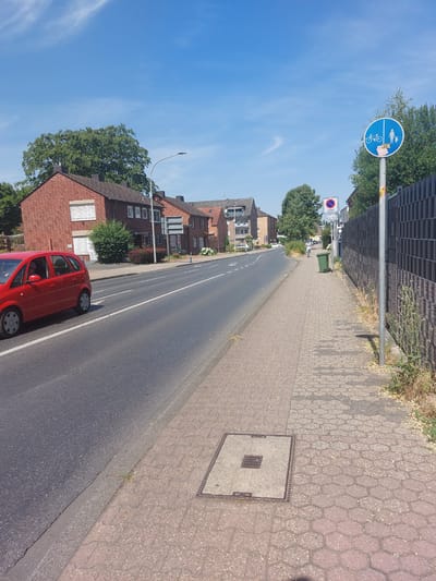The image depicts a street scene in Herzogenrath, Germany. A red car is driving on the road. A sidewalk runs parallel to the road, paved with interlocking concrete blocks. A street sign showing a bicycle and a pedestrian icon is mounted on a pole. Further down the sidewalk, there is a green trash can and a "ZONE" sign with a parking prohibition symbol. Buildings, some made of red brick, line the street. Trees and a black metal fence are also visible. The sky is blue with scattered clouds.