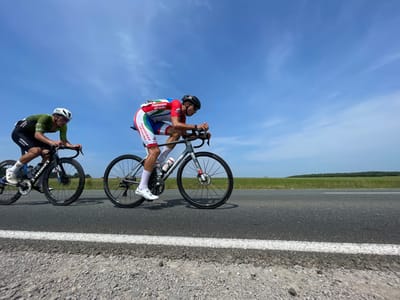 Two male cyclists are racing on a paved road under a bright blue sky with scattered white clouds. The cyclist in the foreground on the right wears a red, white, and blue racing suit with the inscription "Тюменская область" (Tyumen Oblast). He is riding a grey bicycle and is wearing a black helmet and white cycling shoes. The cyclist on the left wears a green and black racing suit and a white helmet. He is riding a black bicycle with "S-WORKS" visible on the frame and is also wearing white cycling shoes. The road has a white line marking and a gravel shoulder. In the background, there is a flat, green landscape with a treeline in the distance. The scene appears to be taking place in or around the village of Perevoznoe, Tyumen Oblast, Russia, based on the inscription on the cyclist's suit.