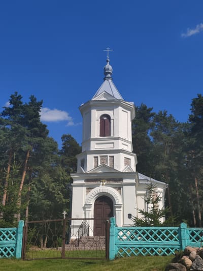 A white, multi-tiered bell tower with a grey, conical roof topped by a cross stands against a bright blue sky with sparse clouds. The tower has a dark wooden door at its base, accessed by a short set of stairs and a black metal gate. The building is surrounded by lush green trees, primarily tall pines with brown trunks. In front of the tower, a turquoise-colored decorative fence with a diamond pattern runs horizontally across the foreground. The ground is covered with green grass, and a few rocks are visible near the fence on the right. The scene appears to be a serene outdoor environment.