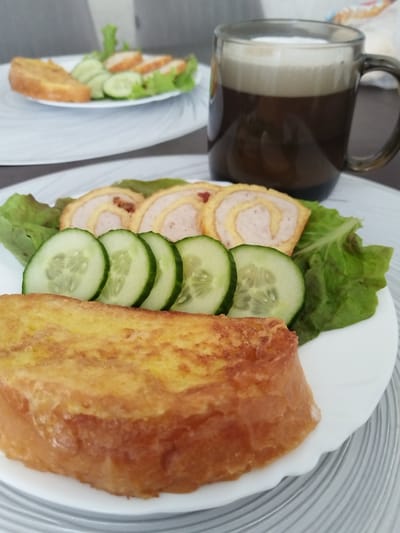 The image shows a close-up of a breakfast setting. In the foreground, a white plate holds a piece of French toast, sliced cucumbers, lettuce, and rolled meat. To the right, a dark glass mug filled with coffee and foam sits on a placemat. In the background, another plate with French toast, cucumbers, lettuce, and sliced meat is visible. The environment appears to be indoors, likely a dining area, with a neutral-colored wall in the background. The scene is well-lit and focuses on the food. The image does not contain any information about the city of Bezlesnyy, Russia.