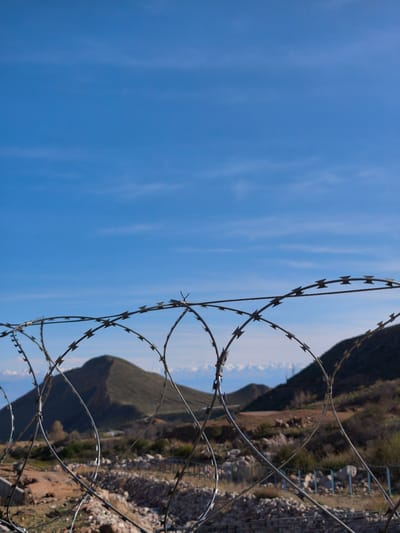 The image depicts a barbed wire fence in the foreground, with a landscape of rolling hills and mountains in the background, all under a clear blue sky. The location is likely in a rural or semi-rural area near Cholpon Ata, Kyrgyzstan, given the terrain and lack of urban structures.
Foreground: A close-up view of a barbed wire fence. The wire is taut and forms sharp barbs, creating a visual barrier. The fence is constructed with multiple strands of wire twisted together, forming a spiral pattern.
Midground: Beyond the fence, there is a dry, rocky landscape with sparse vegetation. Low hills rise in the distance, with larger mountains visible behind them.
Background: A clear blue sky fills the upper portion of the image, with faint wisps of clouds suggesting good weather conditions.