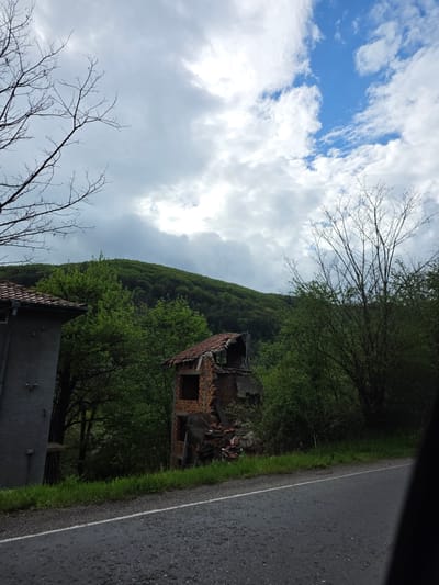 The image shows a ruined brick building with a collapsed roof, located near a road and a green, forested hillside. A grey building is visible in the lower part of the image. The sky is partly cloudy. The location is Gintsi, Bulgaria.