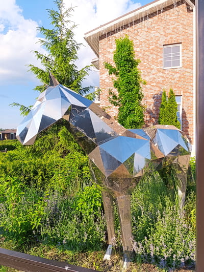 A modern, geometric sculpture of a horse made of polished metal stands in a garden setting. The sculpture is highly reflective, mirroring its surroundings. Behind the sculpture, a brick building with white-framed windows and white icicle-like decorations along the roofline is visible. Various green trees and lush vegetation, including plants with small purple flowers, surround the sculpture. The sky is blue with scattered clouds. The scene appears to be outdoors, possibly in a residential or landscaped area. The specific location is a street in Ivanovskoe, Russia, as indicated by a partial sign visible in the image.
