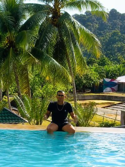A young man is sitting on the edge of a swimming pool with his legs submerged in the turquoise water. He is wearing a black t-shirt, black shorts, sunglasses, and a silver chain. His hands are raised with his thumbs up, indicating a positive gesture. The background features lush green palm trees and dense foliage, suggesting a tropical environment. In the distance, there are buildings with colorful decorations and a hilly, tree-covered landscape. The overall setting appears to be a resort or a private residence in Silaiyajae, Indonesia, characterized by its natural beauty and tranquil atmosphere.