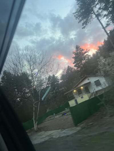 The image shows a sky with gray clouds and a hint of orange light near the horizon. Silhouetted trees are visible in the foreground. A yellow two-story building is slightly visible to the right of the tree line. The image appears to have been taken from inside a vehicle looking through a window. The location, Lukovo, Bulgaria, is not readily discernible from the visual data.