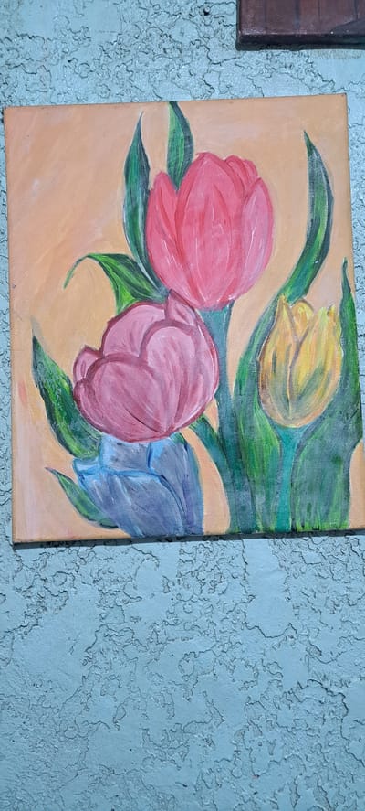 The image shows a close-up, vertical shot of a painting of tulips. The painting is on a light orange background and features three prominent tulips in the foreground: one large pink tulip on the left, a slightly smaller pink tulip in the center, and a blue tulip on the right. There is also a yellow tulip visible in the upper background, partially obscured by green leaves. The tulips are depicted with broad, painterly strokes. The leaves are a vibrant green, adding contrast to the soft colors of the flowers and background. The painting itself appears to be an acrylic or oil on canvas, with a slightly textured surface.

The painting is mounted on a textured wall that appears to be painted a pale, weathered blue. The texture of the wall is rough and uneven, with visible chips and indentations. To the left of the tulip painting, a small portion of another framed artwork with a dark brown frame and dark brown textured content is visible. The overall impression is of artwork displayed in an indoor setting, possibly a home or gallery, against a textured wall. There is no visible text in the image. The lighting suggests it is daytime, with the light source coming from the side, casting subtle shadows on the textured wall.