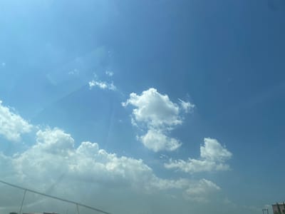 The image captures a bright, expansive blue sky with scattered white clouds, likely taken through a window, possibly from inside a vehicle or building. The primary subject is the sky, dominated by vibrant blue hues.
Numerous cumulus clouds are visible, ranging from small, wispy formations to larger, more defined, fluffy masses. A prominent band of denser white clouds stretches across the upper portion of the frame (when rotated to a landscape orientation). The clouds are brightly lit, suggesting direct sunlight.
Reflections on the glass surface are noticeable, particularly in the lower right and along the top edge, indicating the camera's position. These reflections create faint, indistinct shapes and lines. Glimpses of what appear to be the tops of distant structures or buildings, along with power lines or poles, are visible in the upper left corner through the glass or as reflections. In the top right, faint linear patterns suggest either a road or architectural details.
The scene depicts a calm, clear daytime environment, indicative of fair weather. The bright illumination of the clouds and sky points to a time of day when the sun is high, likely late morning or afternoon. No specific activity or event is taking place, other than the observation of the sky. There is no visible text in the image. Given the provided context, the image captures a sky view from Crescent, Nigeria, though no specific visual cues within the image confirm this location.