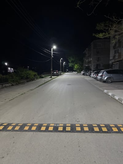 This is a nighttime photograph of a street in Shishmantsi, Bulgaria, viewed from behind a speed bump. The street is paved and illuminated by streetlights. On the right side of the street, several cars are parked parallel to the curb, and an apartment building is visible behind them. The left side of the street features a sidewalk, some greenery, and further streetlights. The sky is dark.