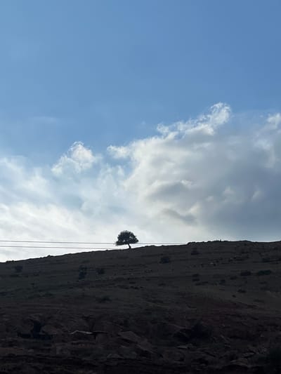 A landscape photograph shows a partly cloudy sky and a hillside. The sky is blue with white clouds. The hillside is dark and rocky, with a small tree growing on it. The scene is likely in a mountainous or rural area. The location is Ait Mazigh, Morocco. The hillside appears to be steep. Power lines are visible in the image.