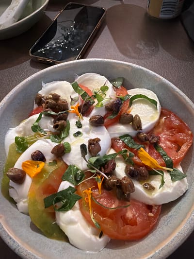 A close-up, overhead shot captures a caprese salad presented in a light blue bowl. The salad is arranged with alternating slices of red tomato and white mozzarella cheese, interspersed with dark olives and fresh green basil leaves. Small yellow flower petals and microgreens add pops of color and texture. The salad appears to be drizzled with olive oil, which glistens under the lighting.

The background is softly blurred, revealing a textured surface, likely a table. To the left of the bowl, a smartphone with a reflective screen is visible, its screen displaying faint reflections of white lights, possibly from overhead lighting or decorative elements. A portion of a bottle with a barcode is also present in the upper left corner. The lighting suggests an indoor setting, possibly a restaurant, with ambient light creating subtle shadows. The overall presentation of the food suggests a meal is being served or enjoyed. There is no indication of people or a specific time of day beyond the artificial lighting. The location context of Platja D'aro, Spain, would imply a Mediterranean dining experience.
