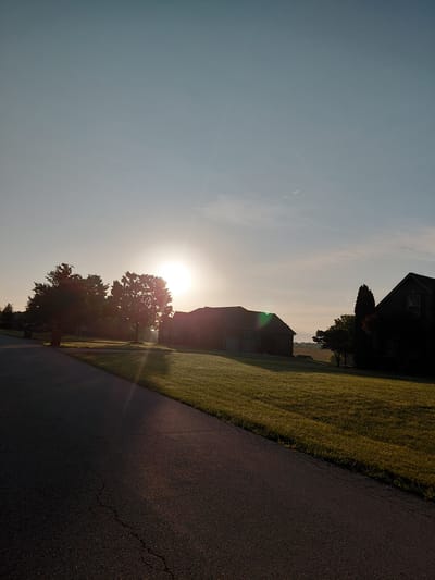The image depicts a rural scene in Paris, United States, at sunrise. The sun is low on the horizon, casting a warm glow and lens flare across the landscape. In the foreground, a dark asphalt road stretches diagonally across the frame, bordered on the left by a lush, green lawn.
Further into the scene, several houses are visible, silhouetted against the bright sky. The architecture suggests suburban or rural dwellings. A prominent feature is a large, leafy tree positioned between two of the houses, its branches illuminated by the rising sun. The sky is a clear, pale blue, gradually transitioning to a brighter white near the sun.
The overall atmosphere is peaceful and serene, characteristic of an early morning in the countryside. The time of day is clearly indicated by the low angle of the sun and the soft lighting. There is no visible text in the image. The scene evokes a sense of calm and the beginning of a new day.