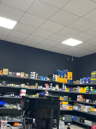 This is an interior shot of a retail store, likely an auto parts shop, in Terpinnia, Ukraine. The camera is oriented vertically, showing shelves stocked with various automotive parts and accessories. The lighting appears to be from overhead fluorescent panels, creating a bright, well-lit environment.
The shelves are filled with boxes of different sizes and colors, many featuring brand names and product information. Prominently visible are boxes labeled "HOLA" and "TADEM," along with other manufacturers. There are also items like shock absorbers, filters, and other mechanical components. The overall impression is one of an organized and well-stocked inventory.
In the foreground, a black retail display unit, possibly a monitor or register, is partially visible, along with some hanging cables and plastic packaging. On the right side of the image, a carton of milk with Ukrainian text ("Молоко 3,2%") indicates the presence of beverages or perhaps a point-of-sale display.
The background shows a dark wall, contrasting with the brightly lit ceiling and the colorful products on the shelves. No people are visible in the image, and the scene appears to be set during business hours. The location cues, such as the "Terpinnia, Ukraine" context and the Ukrainian text on the milk carton, firmly place the setting.