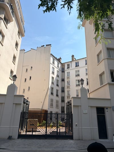 This image depicts a narrow urban courtyard in Courbevoie, France, framed by tall, cream-colored apartment buildings. The perspective is looking upwards, with the sky a clear, bright blue. Lush green foliage from a tree on the left drapes into the frame.

The buildings feature numerous windows with white shutters, many of which are open, suggesting a warm time of day. Architectural details include balconies with decorative metal railings and prominent chimney stacks atop the buildings. A classic black street lamp with an ornate design stands out against the pale facades, positioned on a white column.

In the foreground, a black metal gate with a geometric pattern guards an entrance to a courtyard area, where a few plants are visible. The ground in the foreground is paved with light-colored rectangular stones. The overall impression is one of a well-maintained, somewhat classic European residential area. The time of day appears to be late morning or early afternoon, given the bright sunlight casting shadows. No people are visible in the frame, and there is no discernible text in the image.